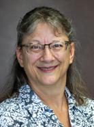 A woman with glasses, gray hair, and wearing a patterned shirt smiles in a headshot against a plain background.