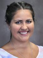 A smiling person with dark hair pulled back, wearing a light-colored top and a necklace, against a neutral background.